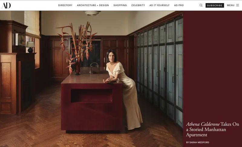 Architectural Digest desktop story page for “Athena Calderone Takes On a Storied Manhattan Apartment”, a photo of a woman in a cream dress leaning on a red kitchen island beside a burgundy title panel